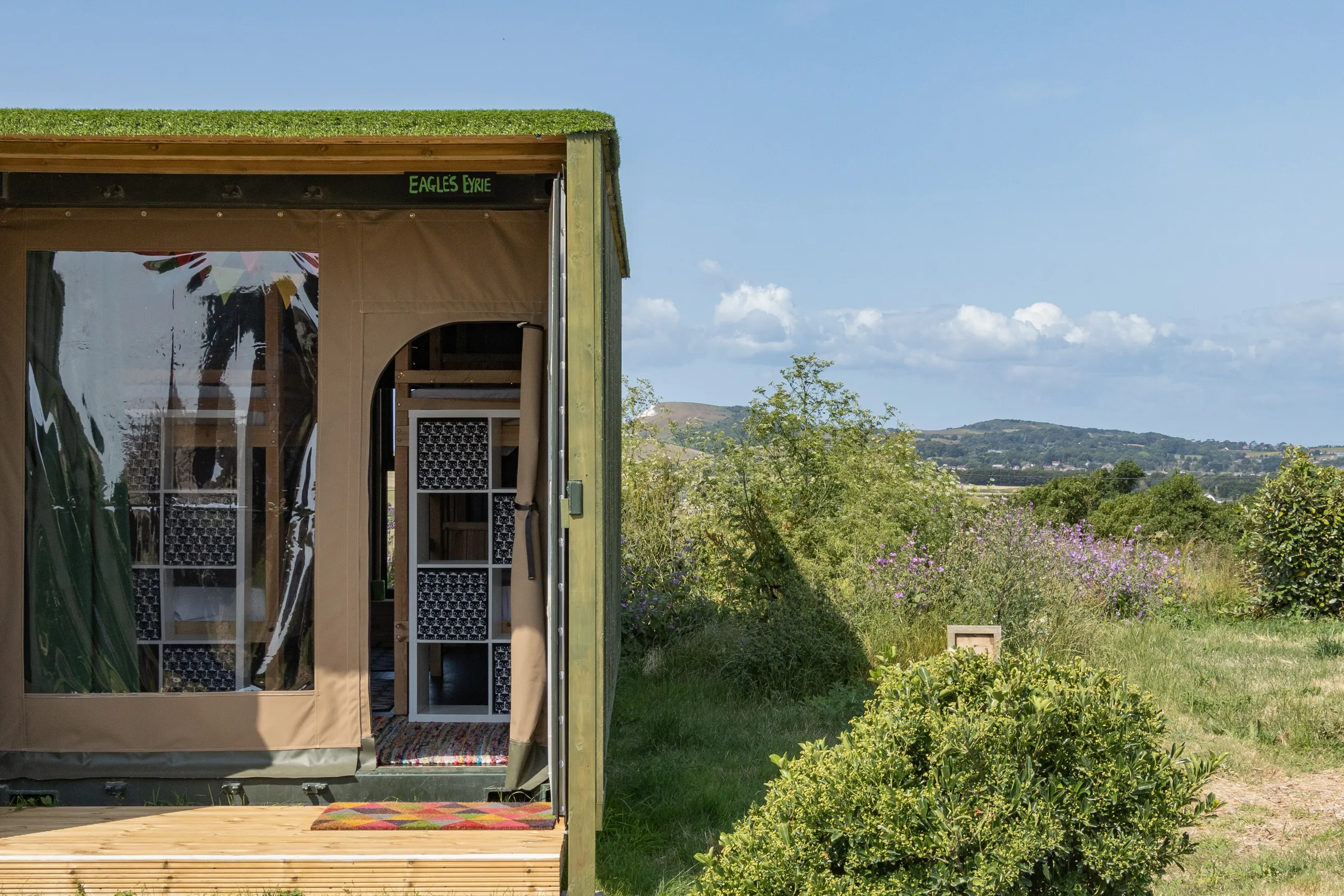 Exterior of an eco-friendly glamping container with an open canvas doorway, overlooking wildflower meadows and distant hills under a blue sky.
