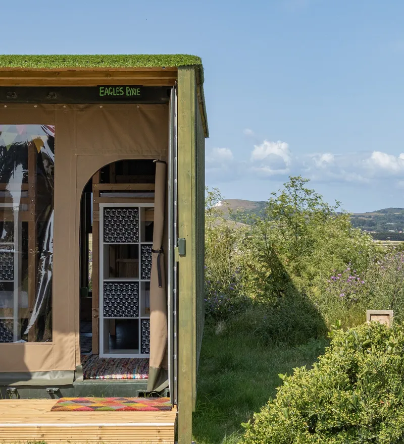 Exterior of an eco-friendly glamping container with an open canvas doorway, overlooking wildflower meadows and distant hills under a blue sky.