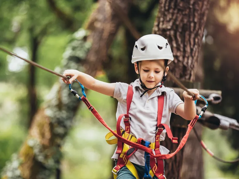 Young child wearing a white helmet and safety harness on a rope course in a forest