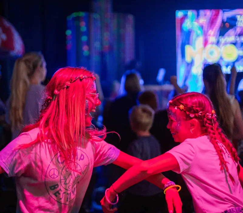 Two girls dancing under bright pink and blue stage lights at a lively concert.