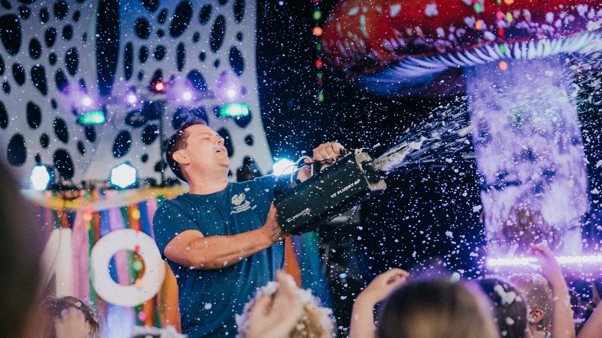 Man operates a foam machine at a colourful indoor party, spraying foam over a crowd beneath stage lights and a giant mushroom decoration