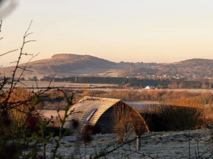 Frosty countryside landscape with a curved wooden barn in the foreground and hills in the distance at sunrise