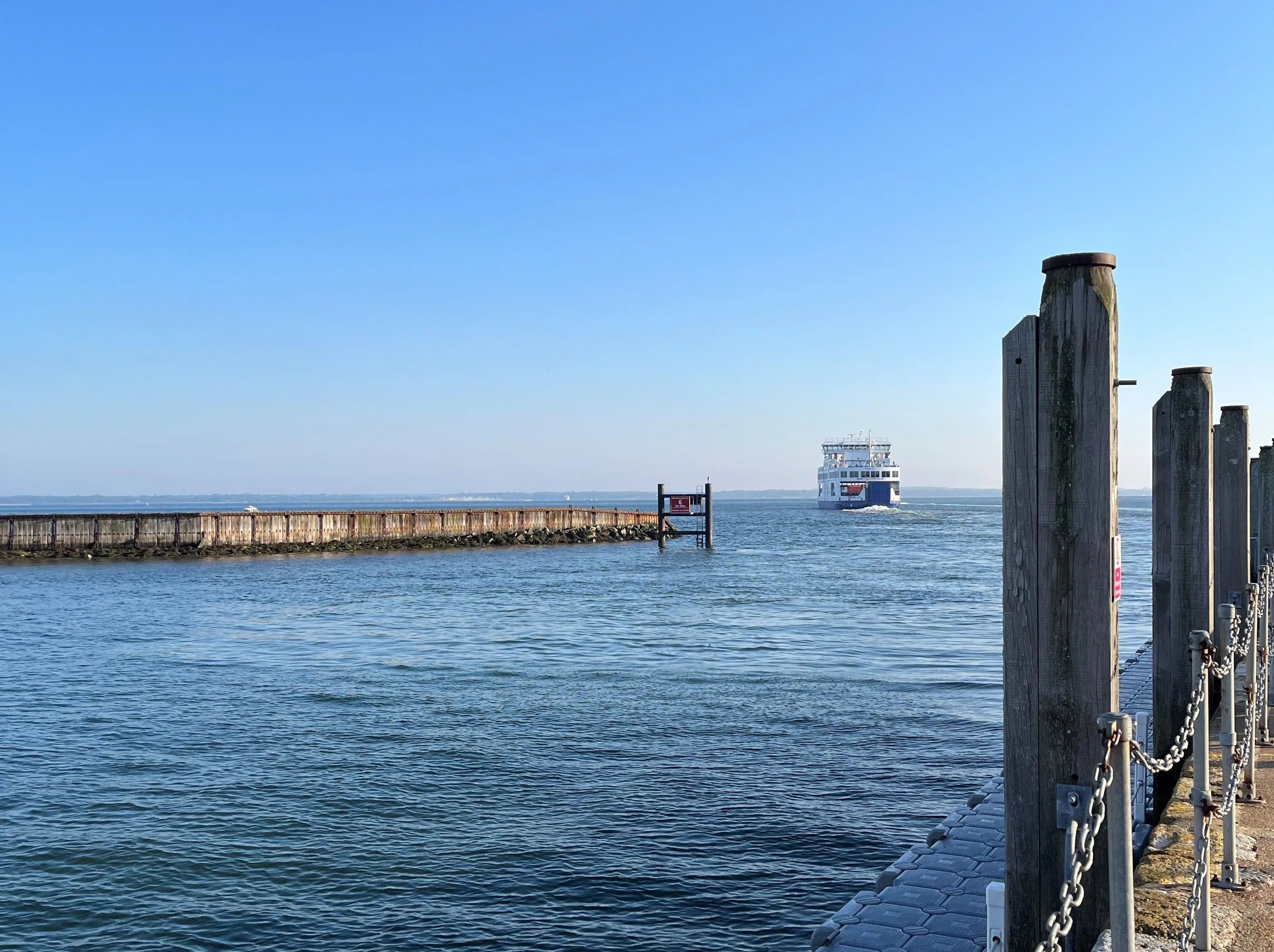 Ferry sailing across calm water near a pier with wooden posts and chain railing under a clear blue sky