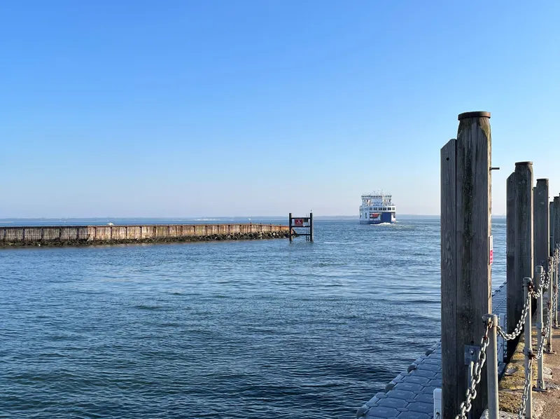 Ferry sailing across calm water near a pier with wooden posts and chain railing under a clear blue sky