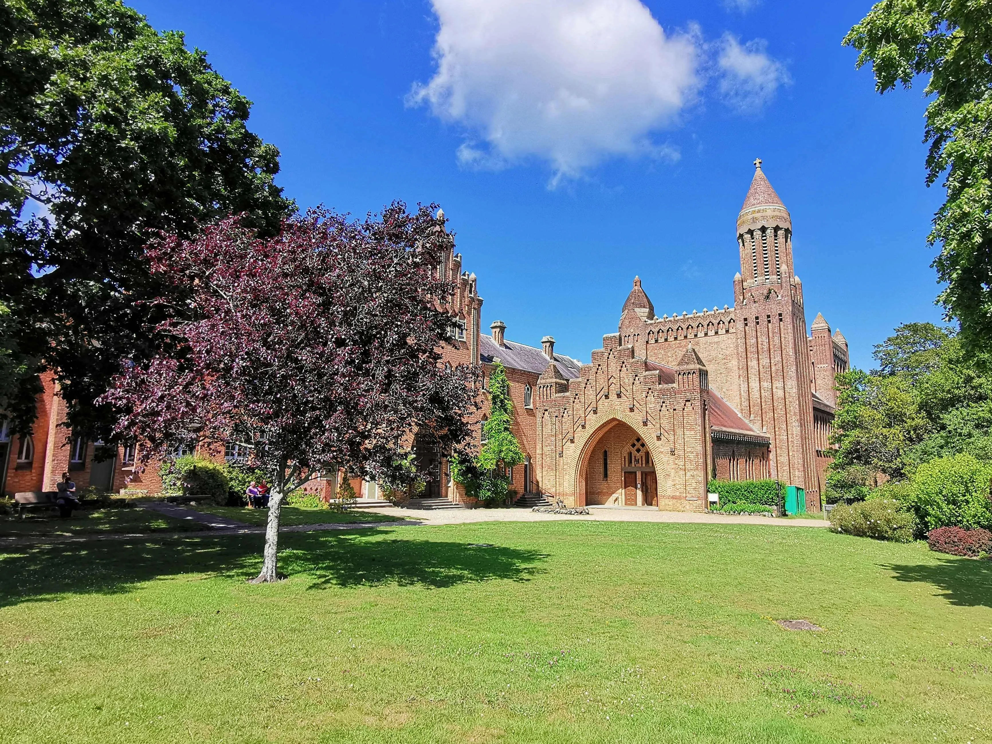 Red-brick historic building with a tower and arched entrance, viewed across a green lawn under a blue sky