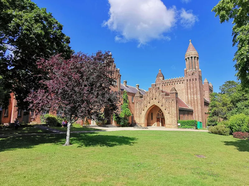 Red-brick historic building with a tower and arched entrance, viewed across a green lawn under a blue sky