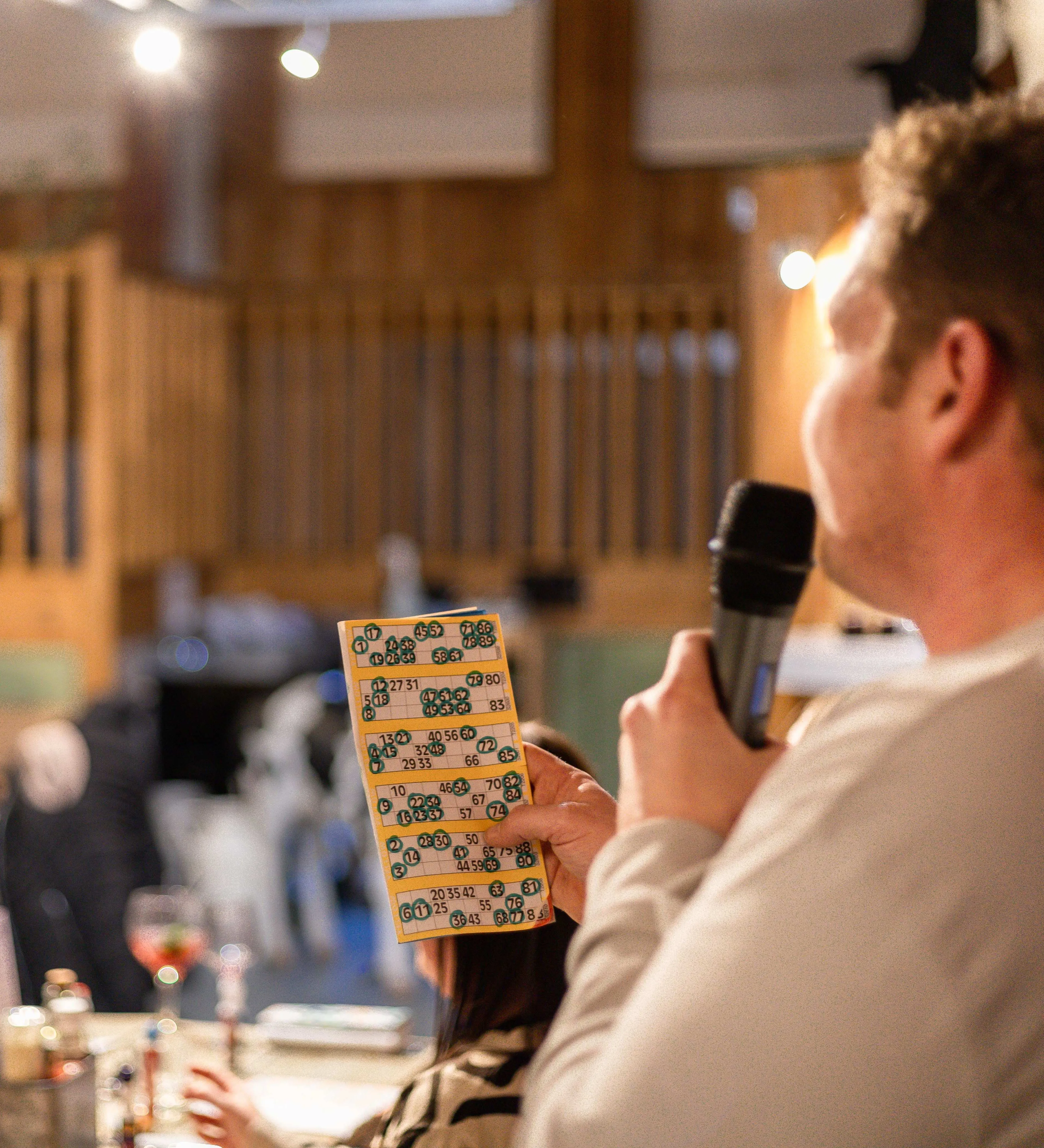 Caller with a microphone holds a bingo card in a warm, rustic pub as players sit at tables in the background.
