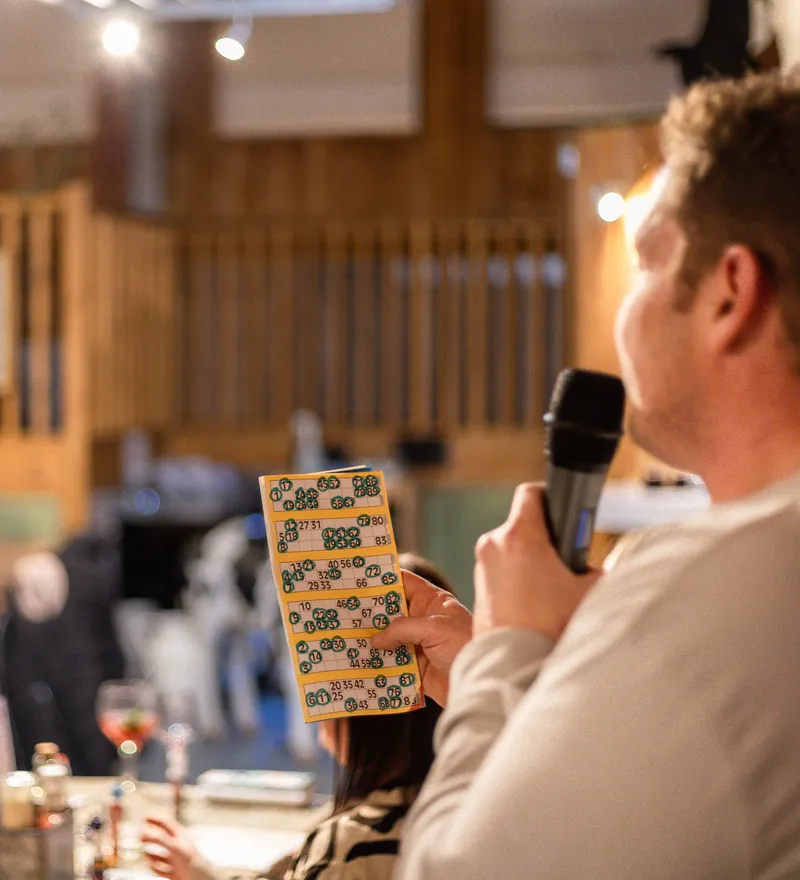 Caller with a microphone holds a bingo card in a warm, rustic pub as players sit at tables in the background.