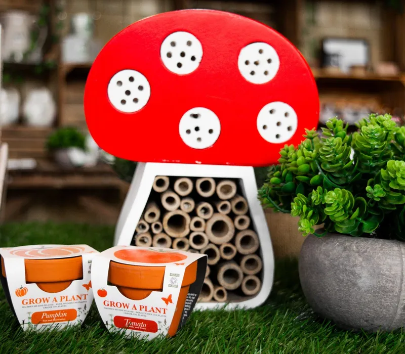 Red mushroom-shaped insect hotel with bamboo tubes beside potted green plant and grow-a-plant kits