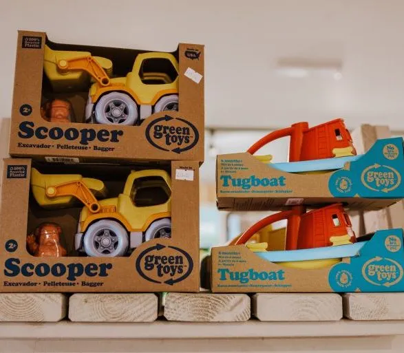 Stacked Green Toys boxes containing a yellow toy digger and tugboats on a shop shelf