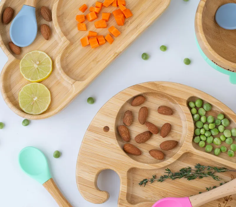 Bamboo baby plates and bowl with colourful silicone-tipped spoons, peas, almonds and sliced lime on a white surface