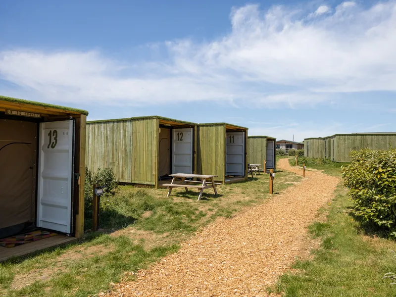 Row of wooden glamping cabins made from shipping containers along a woodchip path under a blue sky