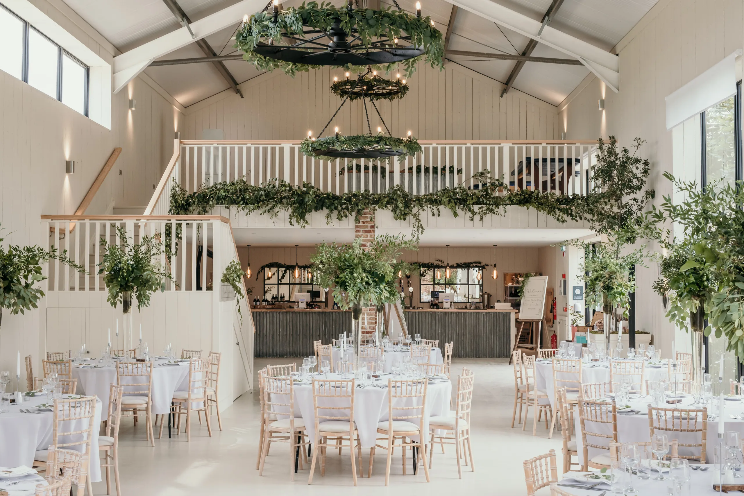 Bright barn wedding reception with round tables, wooden chairs and greenery chandeliers