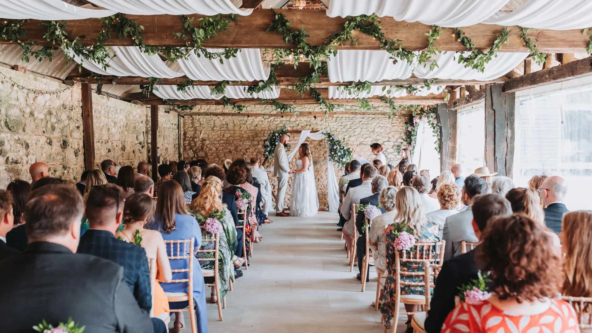 Wedding ceremony in a rustic barn with guests seated in rows and the couple standing at the front beneath floral decorations