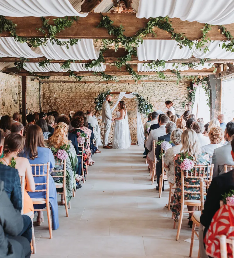 Wedding ceremony in a rustic barn with guests seated in rows and the couple standing at the front beneath floral decorations