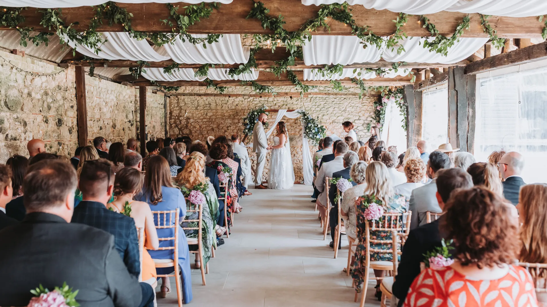Wedding ceremony in a rustic barn with guests seated in rows and the couple standing at the front beneath floral decorations