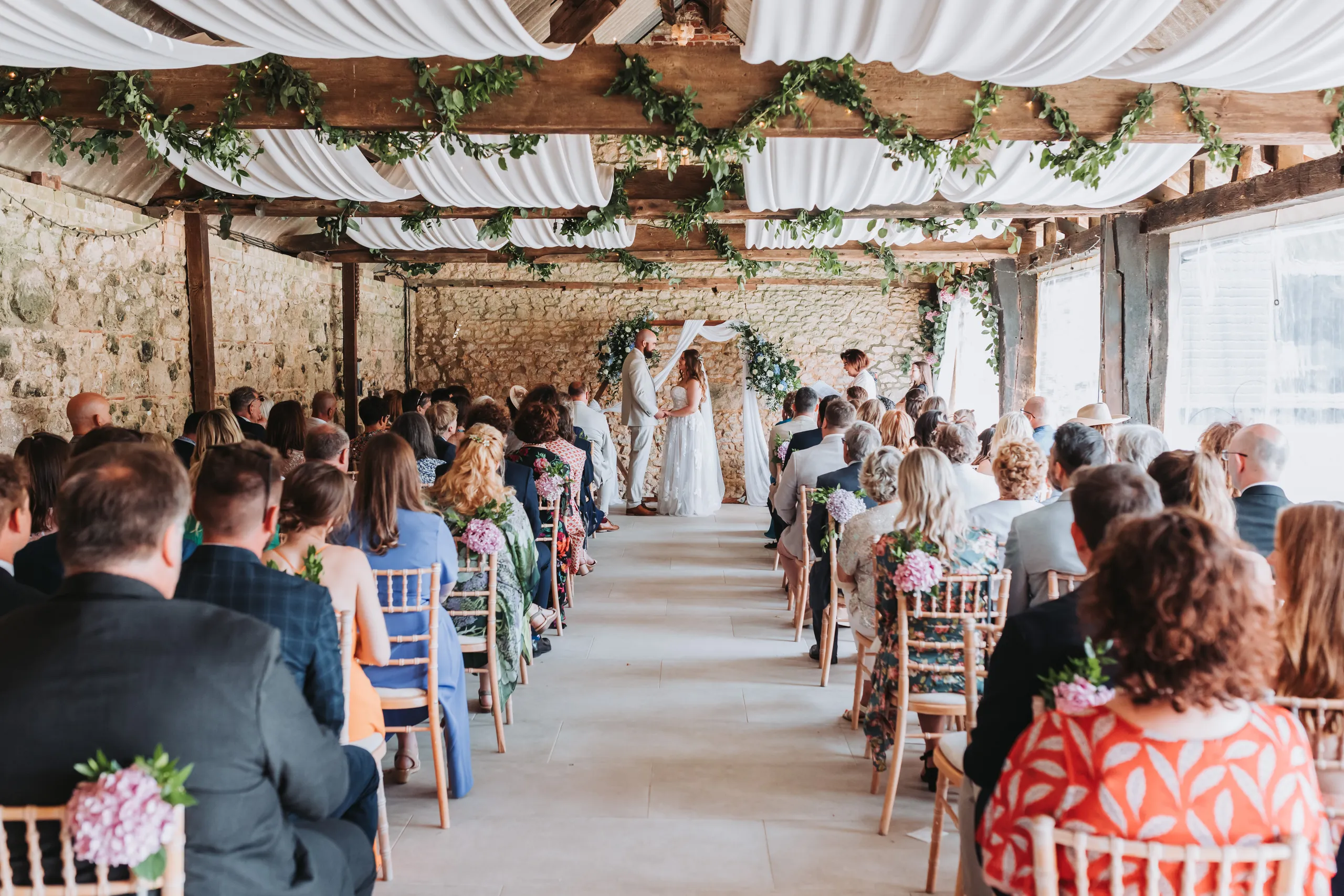 Wedding ceremony in a rustic barn with guests seated in rows and the couple standing at the front beneath floral decorations