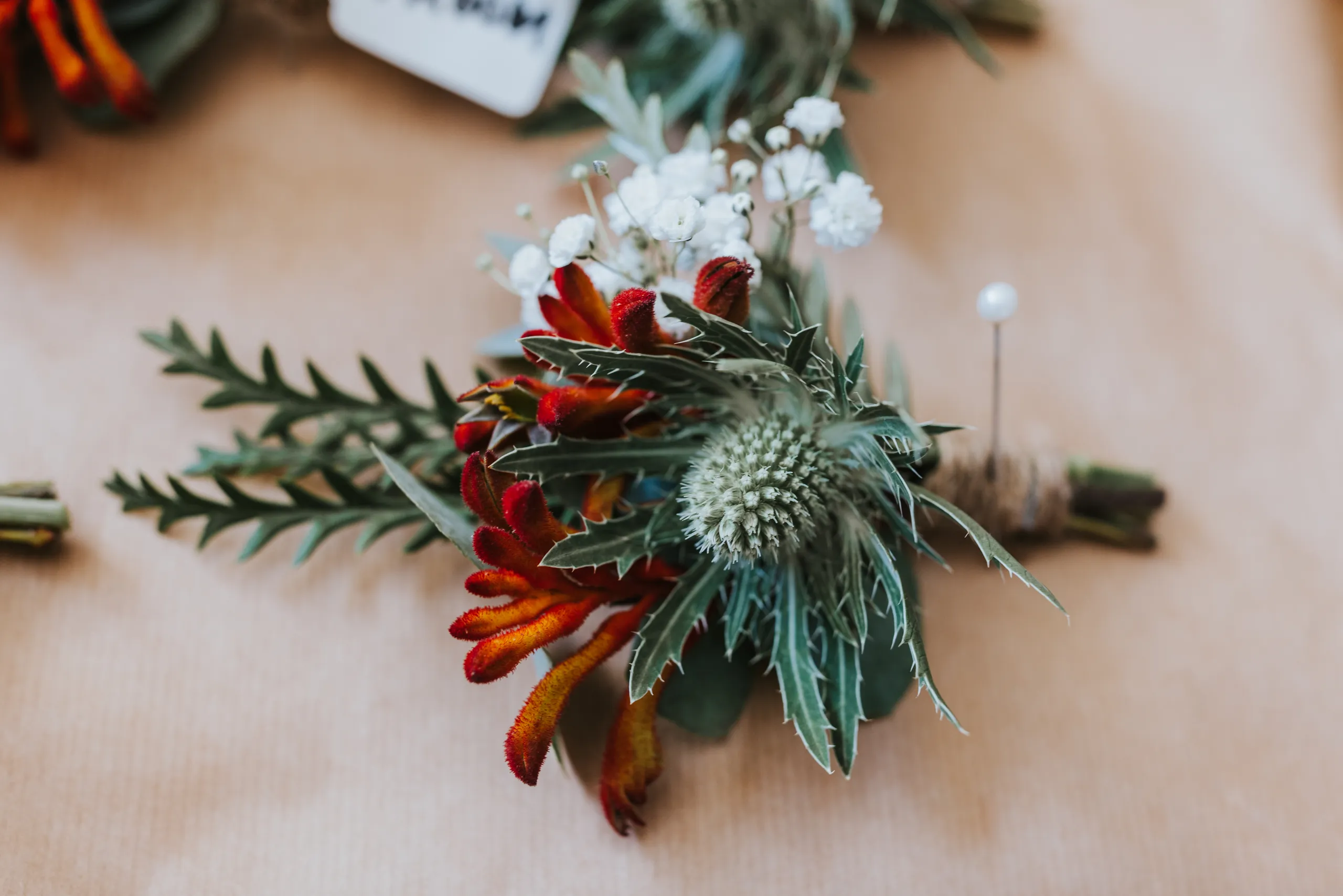 Close-up of a rustic boutonniere with red and orange kangaroo paw, white baby's breath, and grey-green foliage on a wooden surface