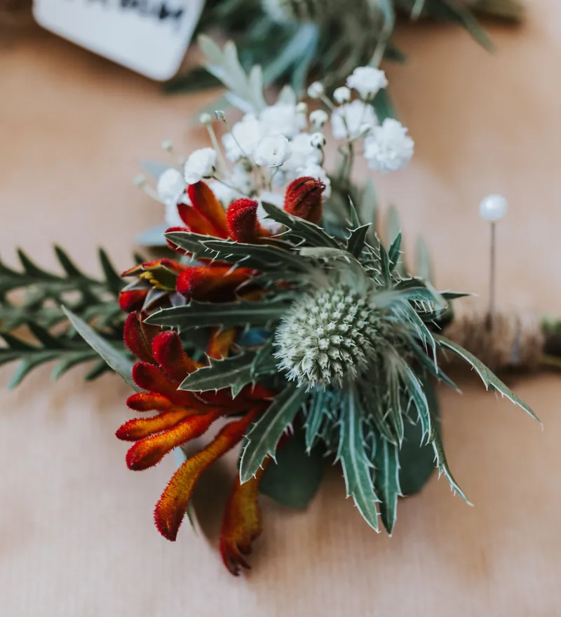 Close-up of a rustic boutonniere with red and orange kangaroo paw, white baby's breath, and grey-green foliage on a wooden surface