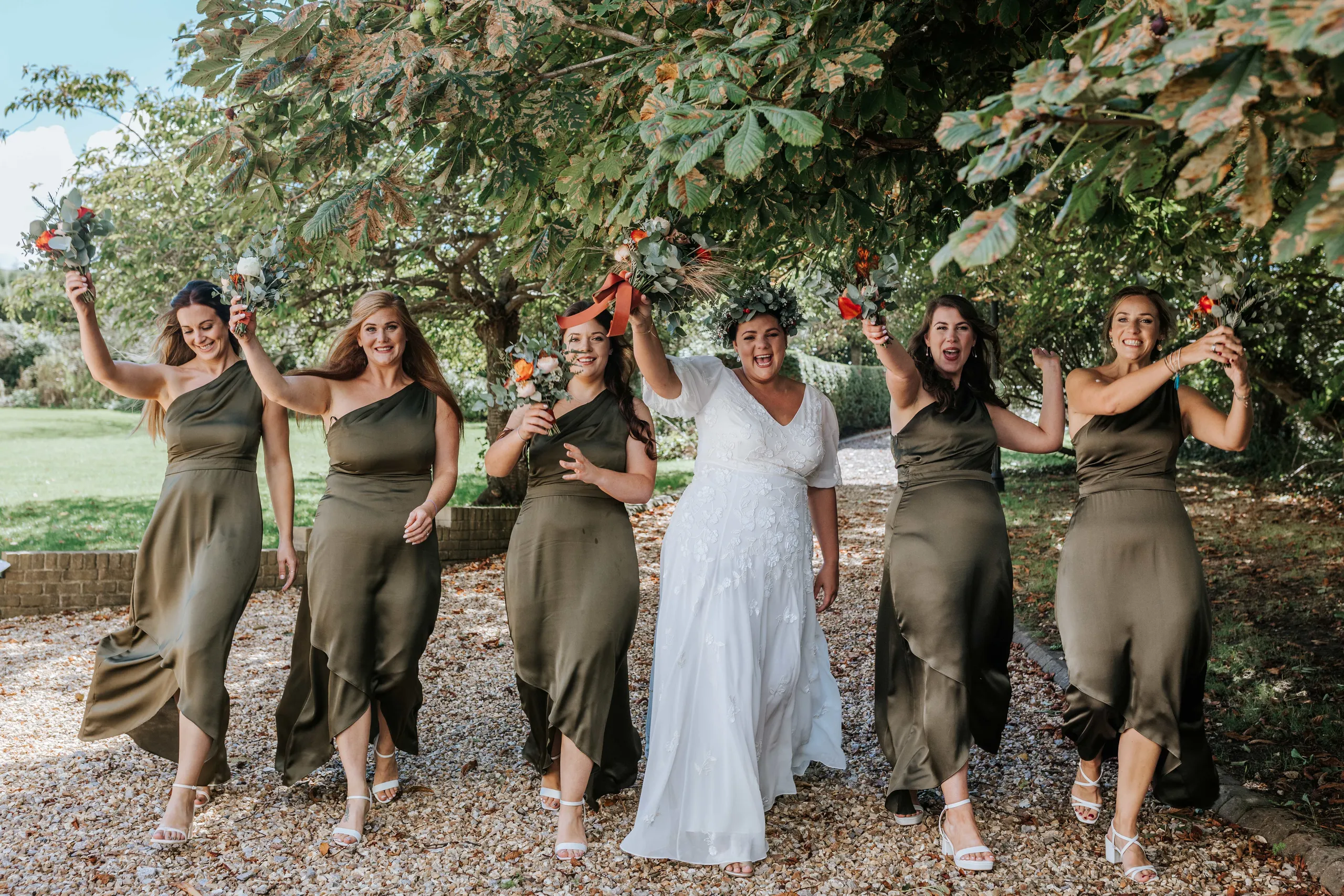 Bride and bridesmaids in olive dresses walking under trees, holding bouquets aloft