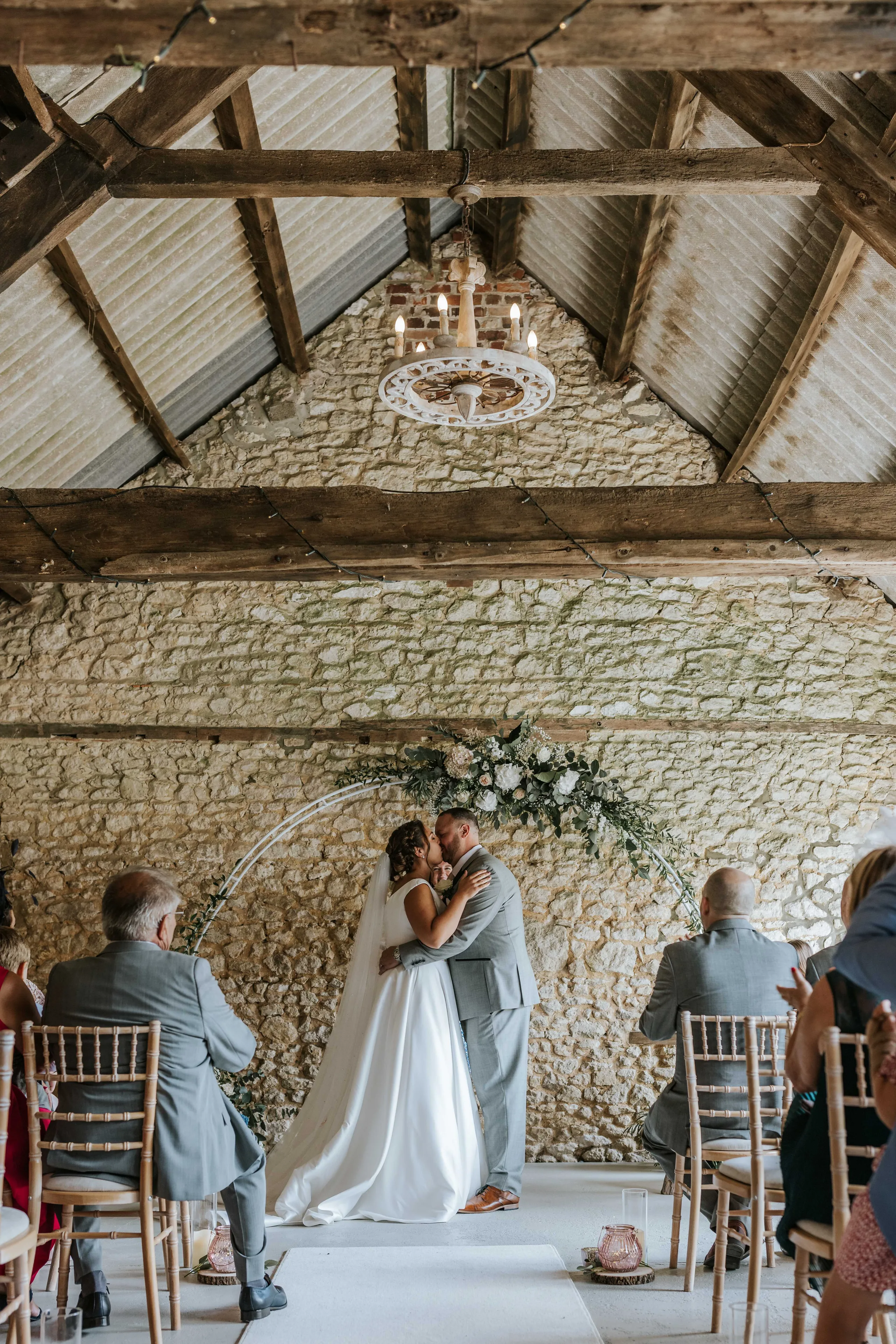 Bride and groom sharing a kiss beneath a floral arch in a rustic stone barn ceremony