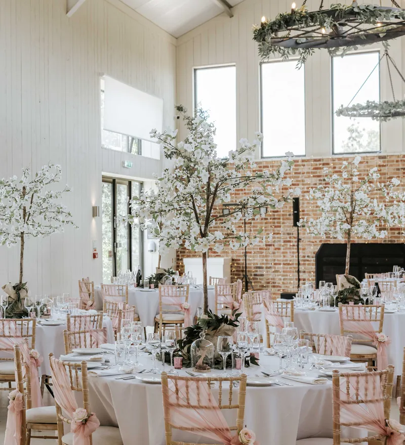 Elegant wedding reception tables in a bright barn venue with blossom centrepieces and pink chair sashes