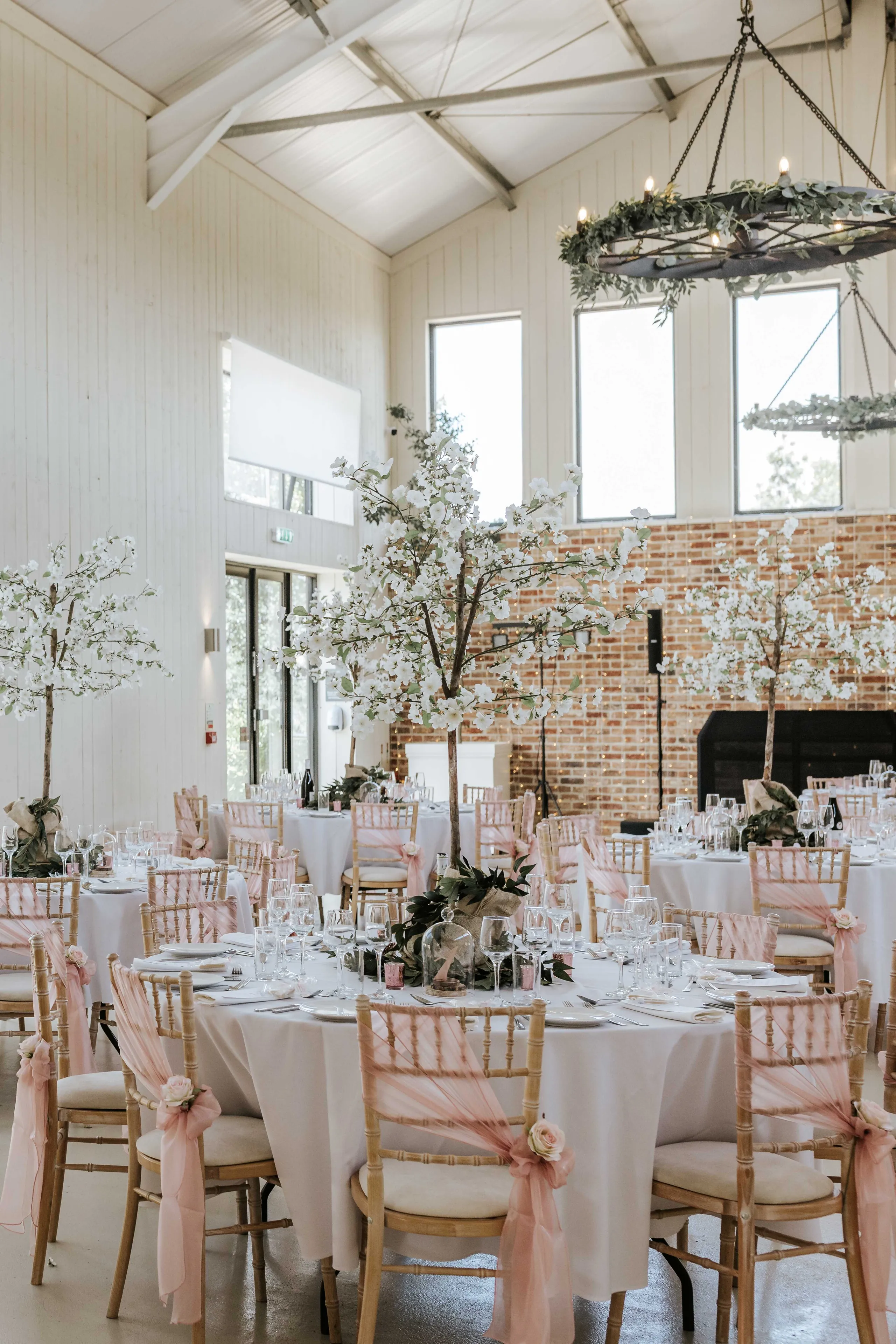 Elegant wedding reception tables in a bright barn venue with blossom centrepieces and pink chair sashes