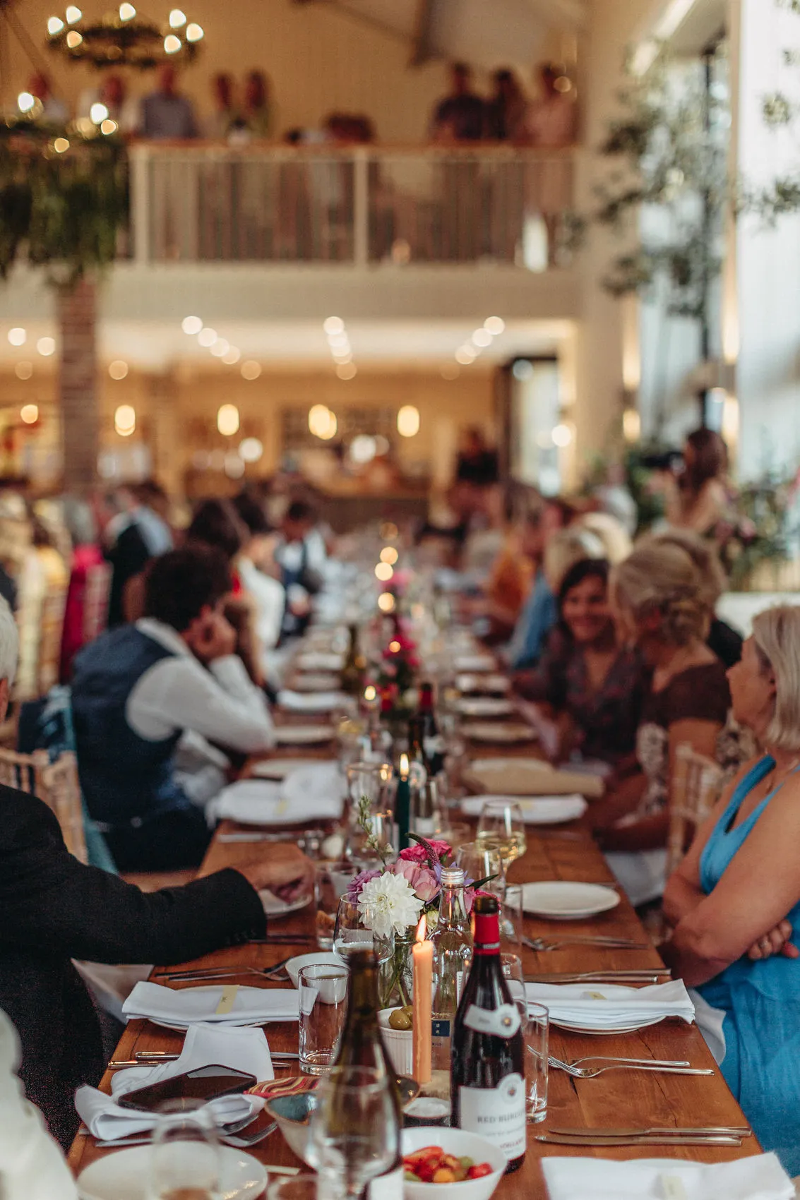 Guests seated at a long wooden dining table with candles, wine bottles and flowers in a bright reception hall
