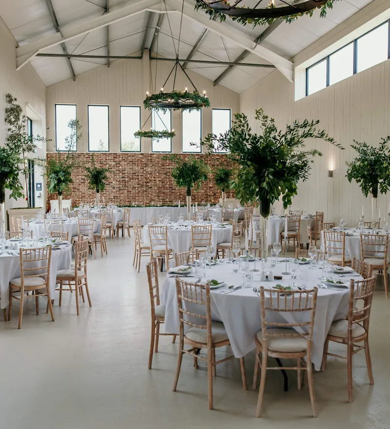 Bright barn wedding reception with round tables, white linens and greenery centrepieces