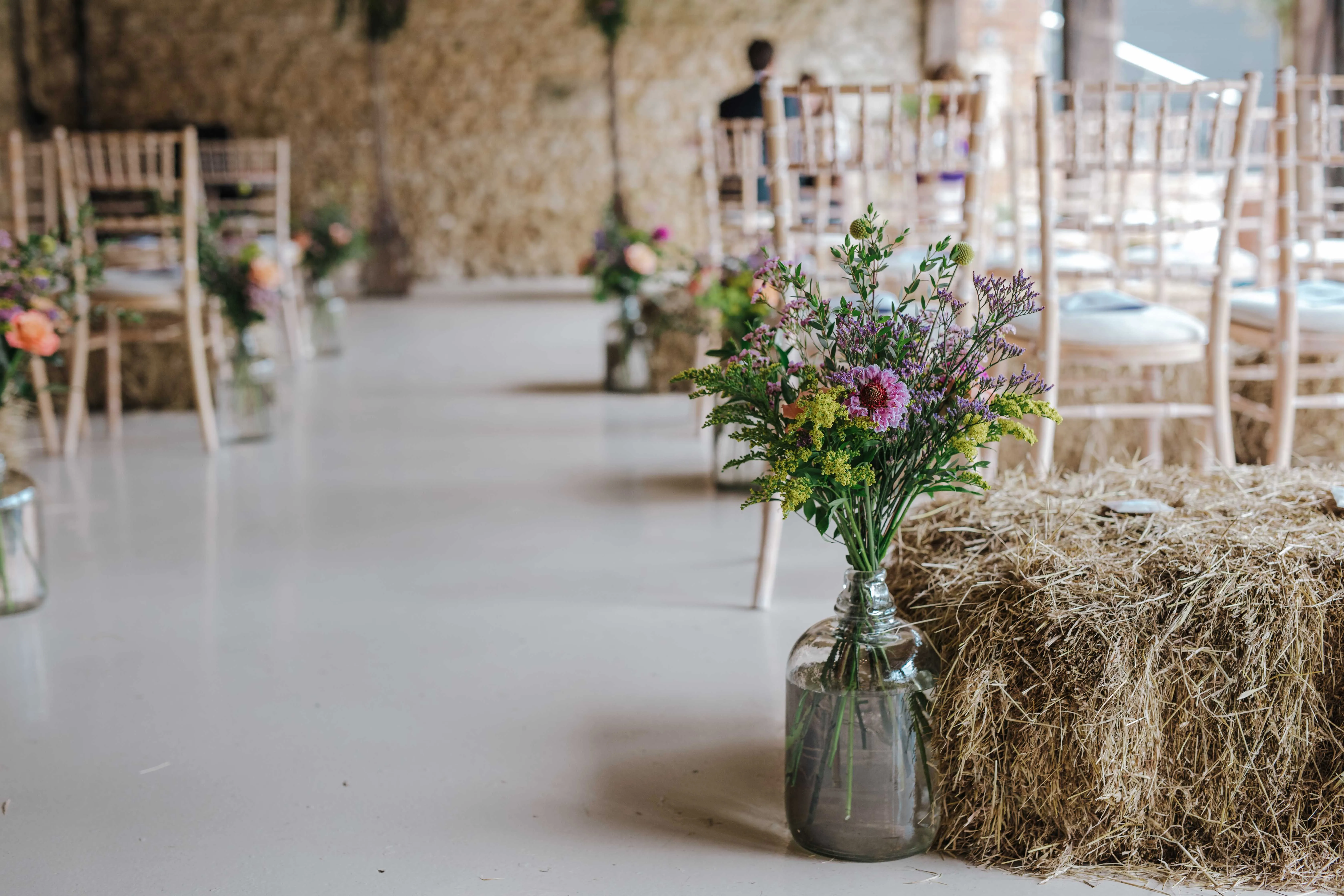 Wildflower bouquet in a glass jar beside a hay bale in a rustic wedding venue with wooden chairs
