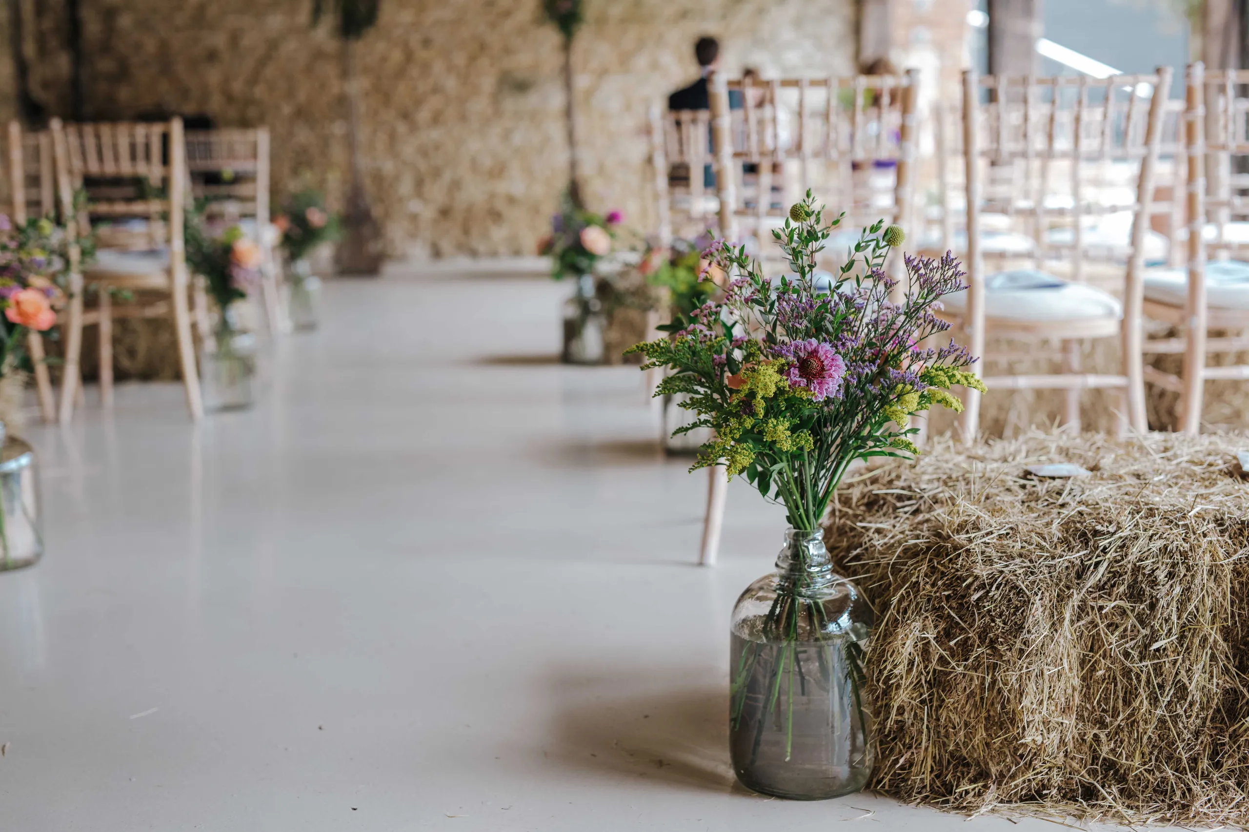 Wildflower bouquet in a glass jar beside a hay bale in a rustic wedding venue with wooden chairs