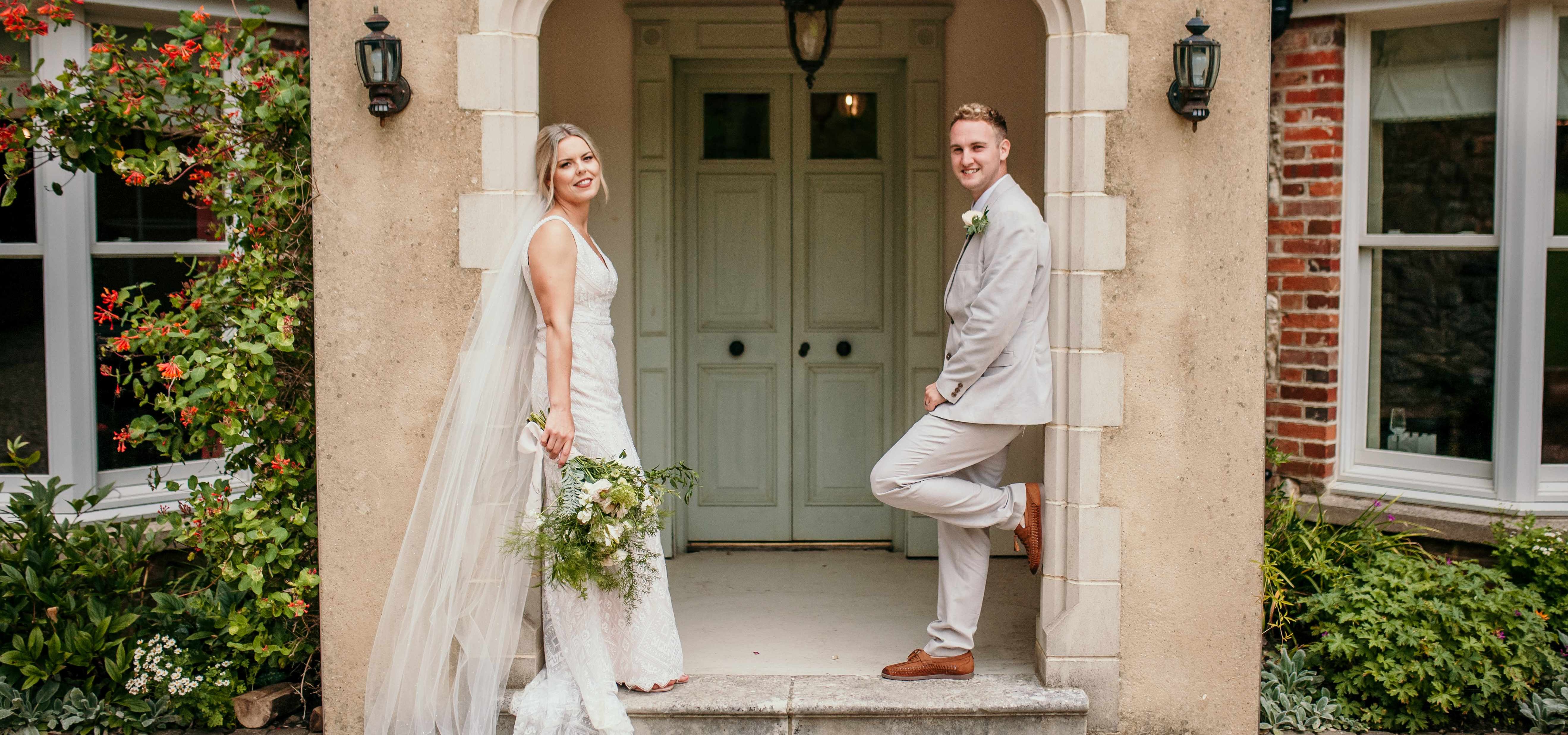Bride in a white lace gown holding a bouquet stands by an arched doorway while the groom in a light suit leans against the entrance of a country house.