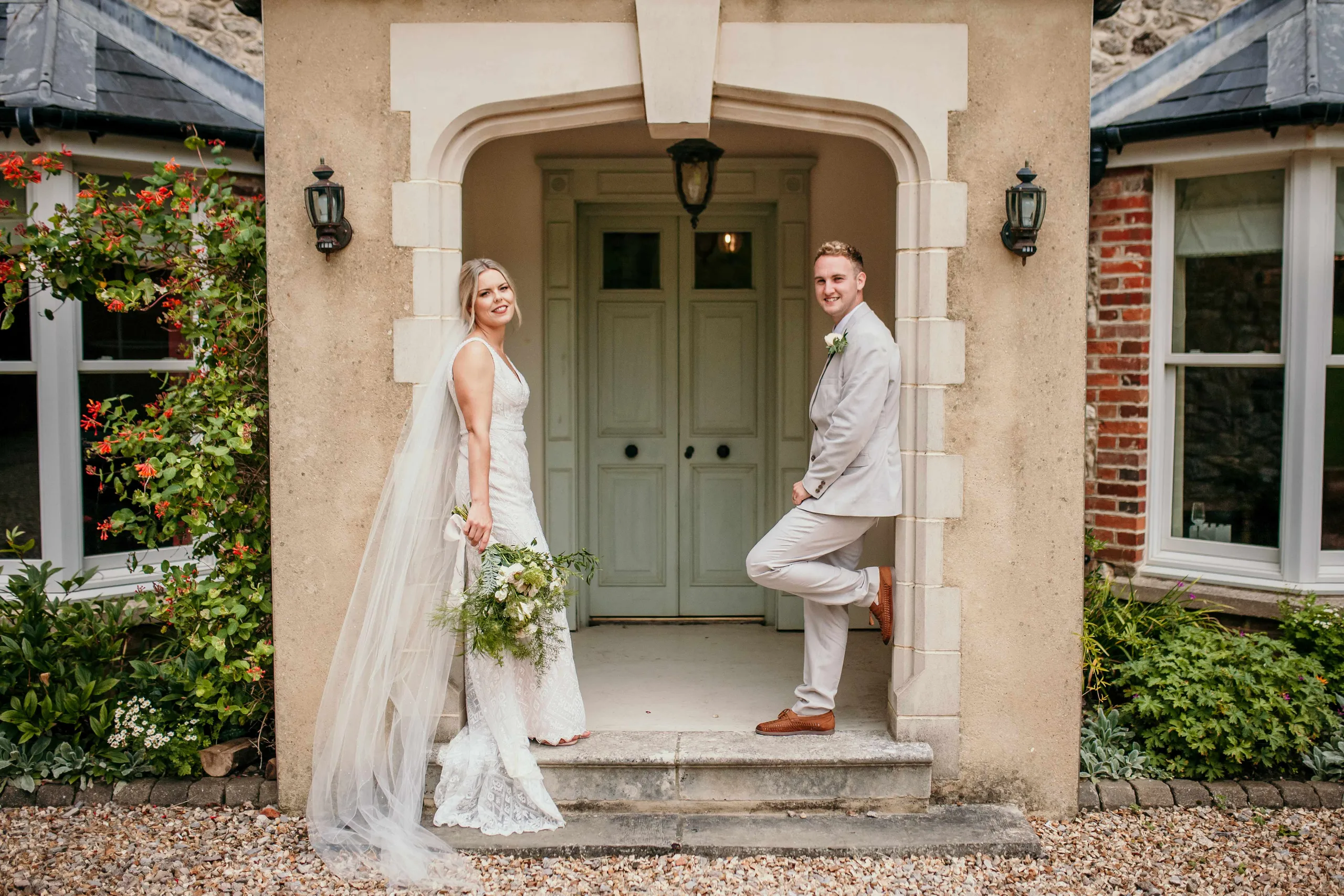 Bride in a white lace gown holding a bouquet stands by an arched doorway while the groom in a light suit leans against the entrance of a country house.