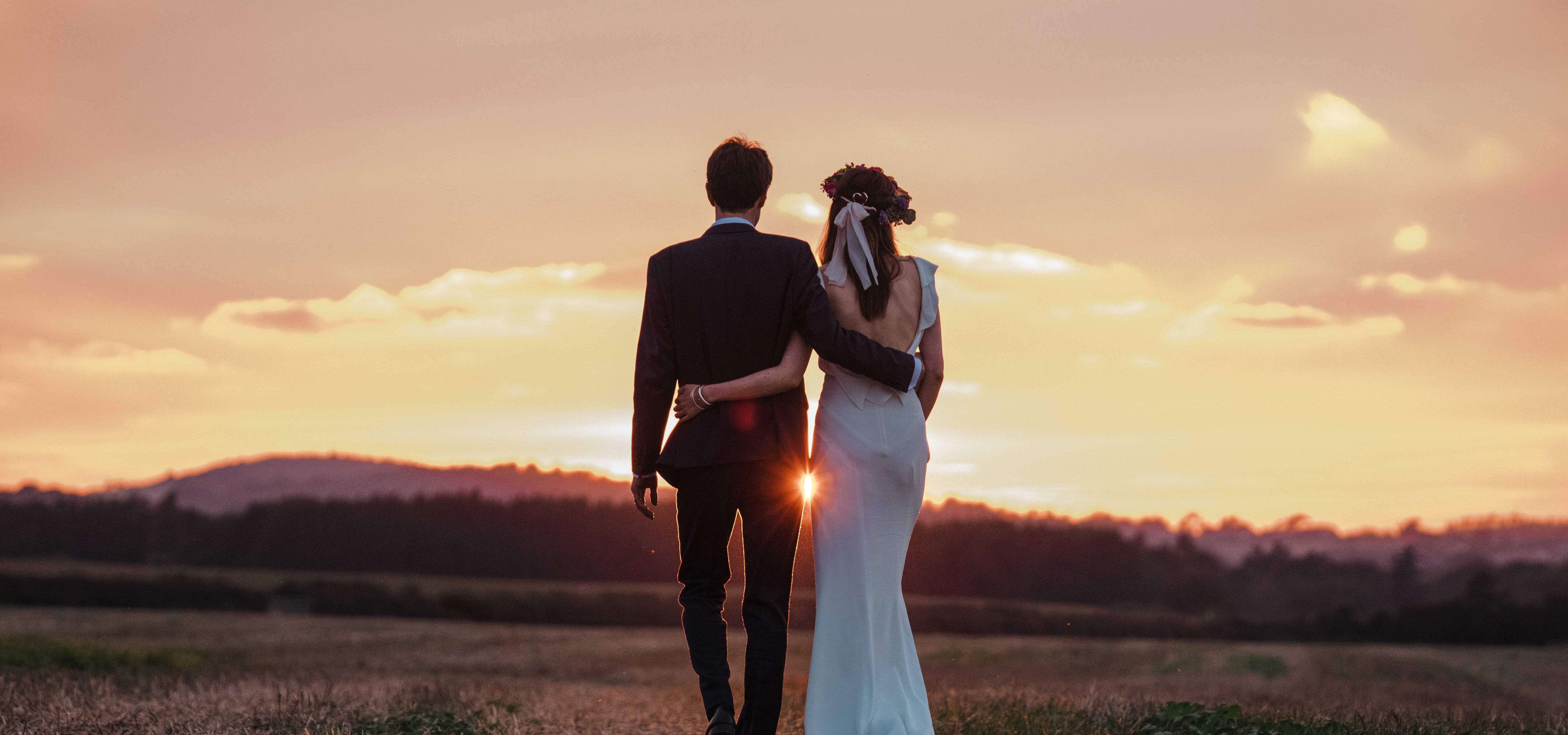 Couple in wedding attire walking arm in arm through a field at sunset