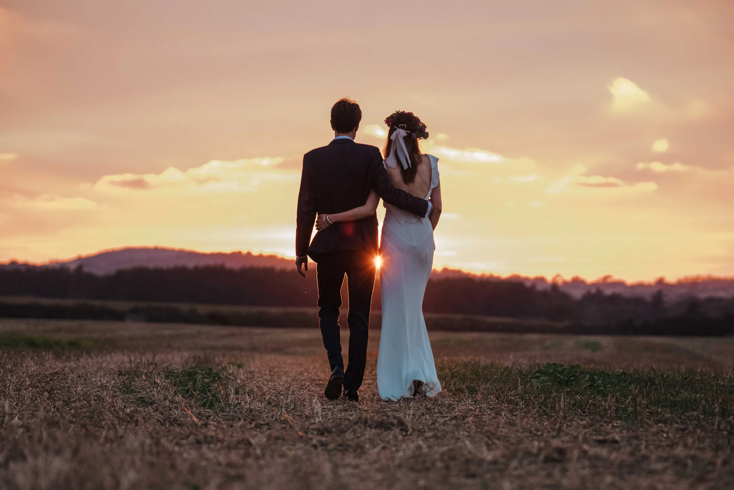Couple in wedding attire walking arm in arm through a field at sunset