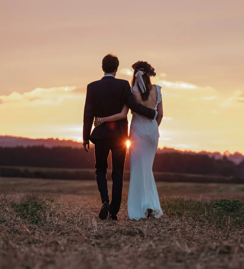 Couple in wedding attire walking arm in arm through a field at sunset