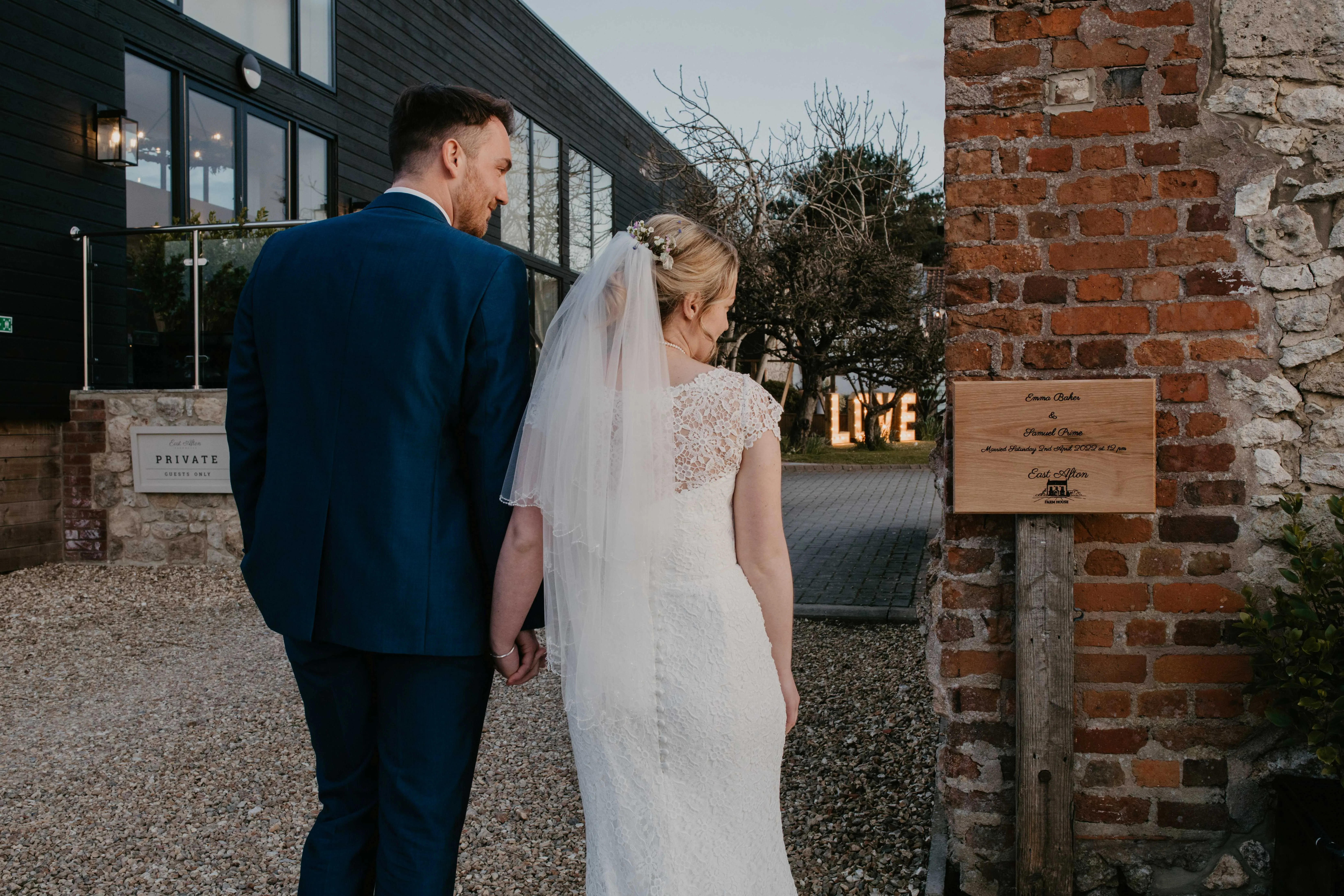 Bride and groom holding hands outside a wedding venue beside a brick wall and wooden sign, with illuminated LOVE letters in the background