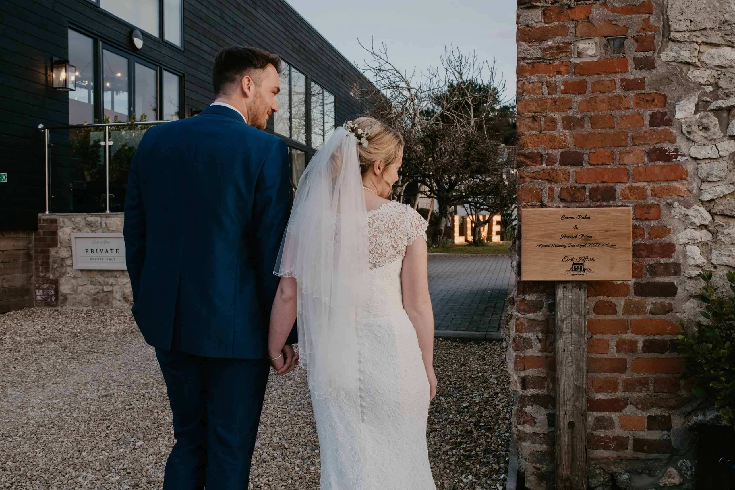Bride and groom holding hands outside a wedding venue beside a brick wall and wooden sign, with illuminated LOVE letters in the background
