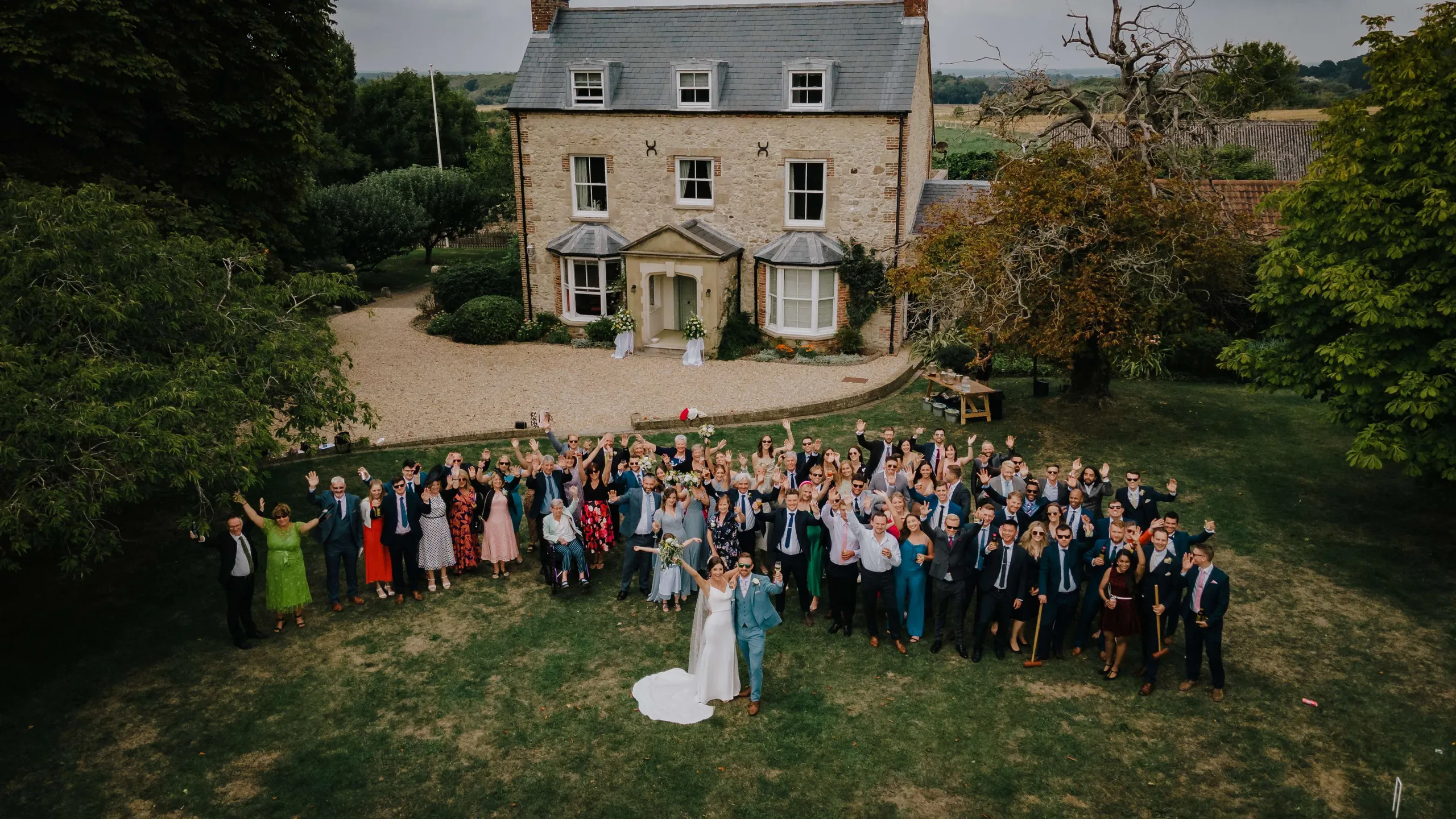 Aerial view of a wedding group on a lawn in front of a stone country house, with the bride and groom posing at the front and guests waving.