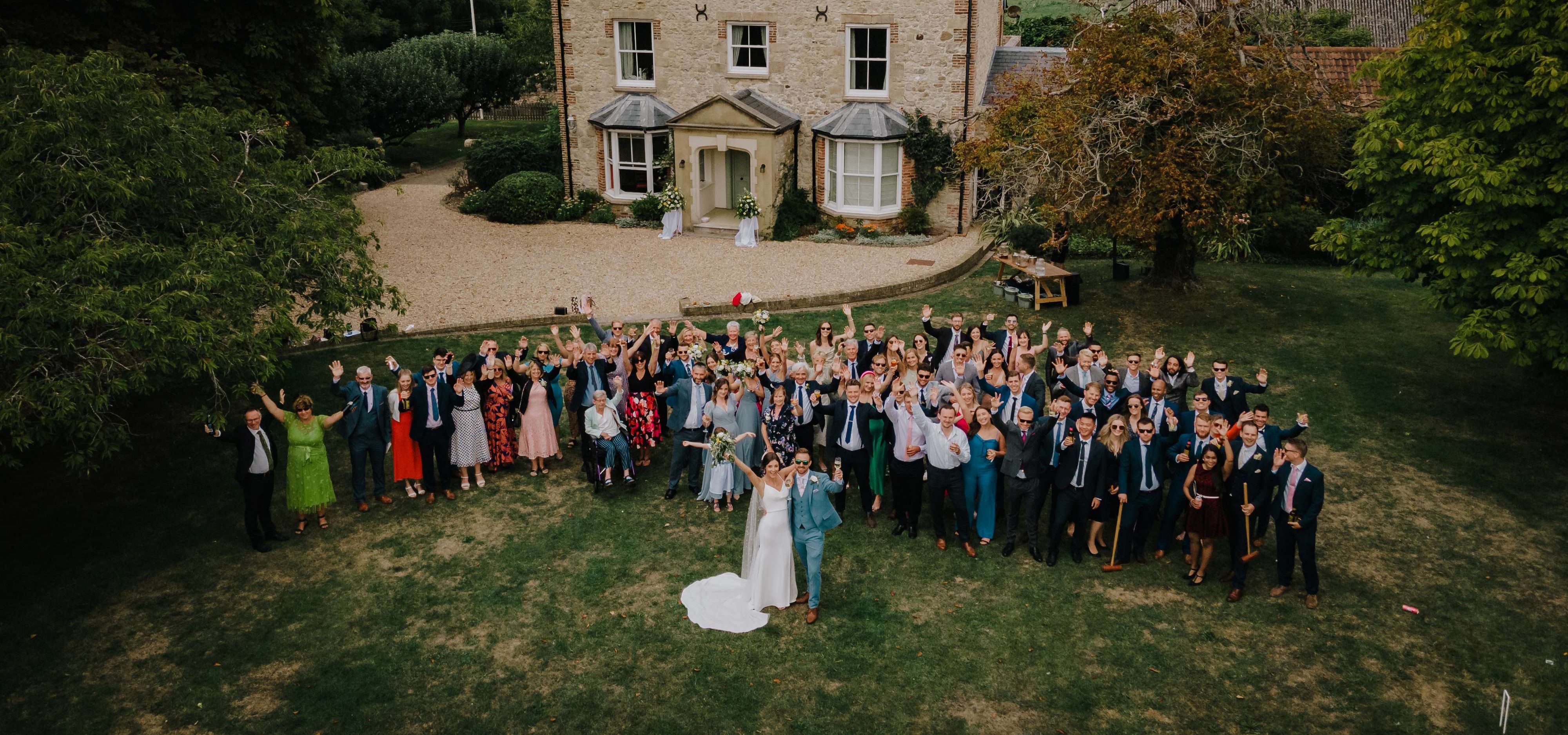 Aerial view of a wedding group on a lawn in front of a stone country house, with the bride and groom posing at the front and guests waving.