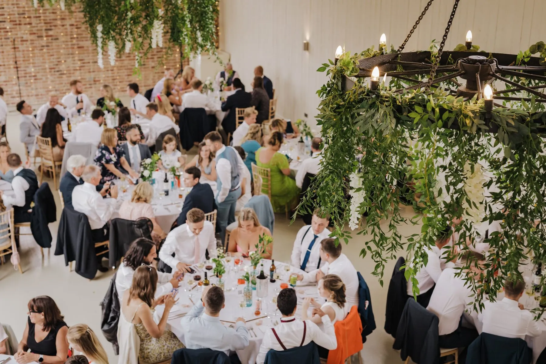Wedding reception guests seated at long tables beneath a greenery-wrapped chandelier