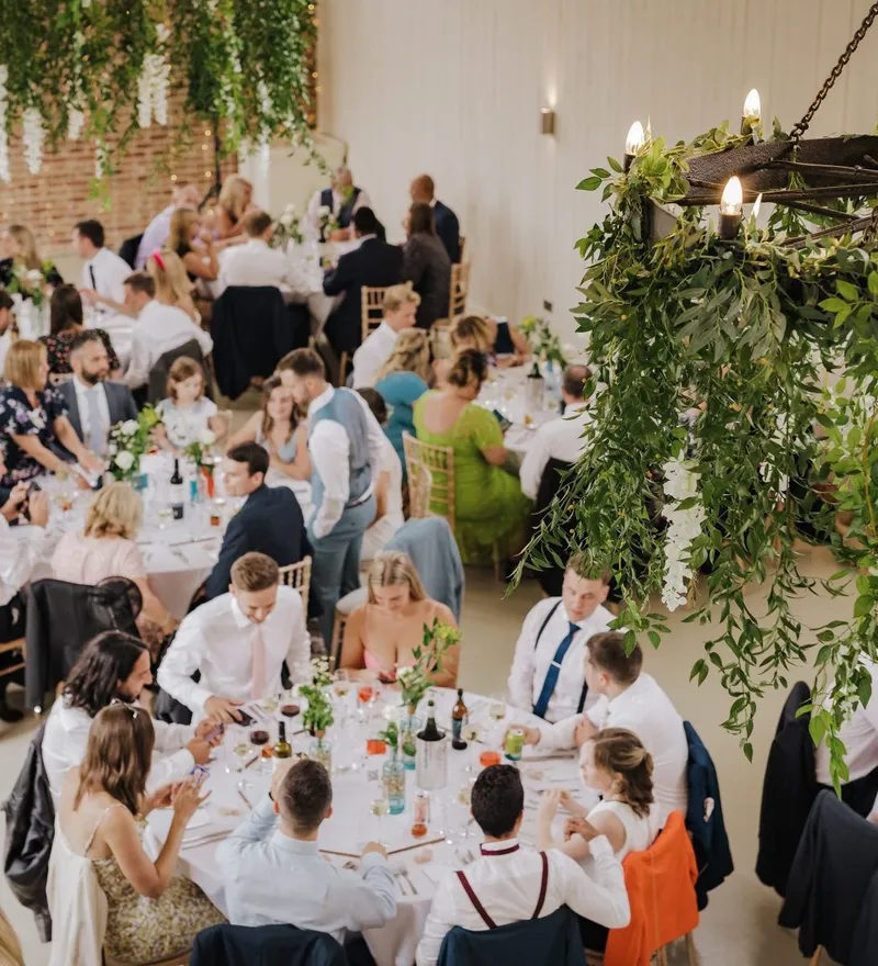 Wedding reception guests seated at long tables beneath a greenery-wrapped chandelier