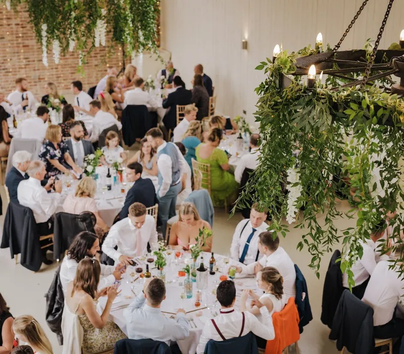 Wedding reception guests seated at long tables beneath a greenery-wrapped chandelier