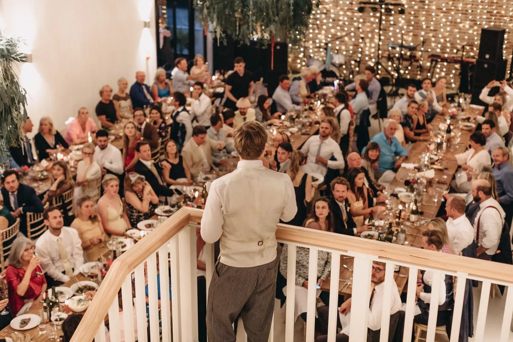 Man giving a speech from a staircase to guests seated at long tables in a warmly lit reception hall