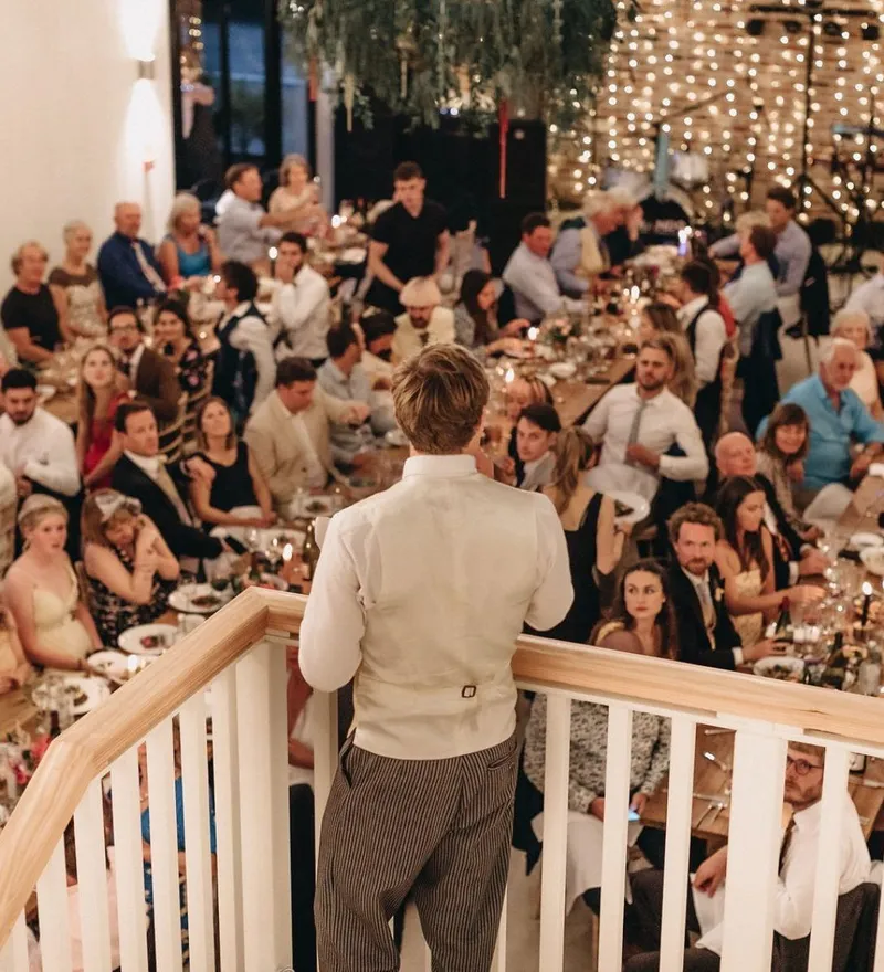 Man giving a speech from a staircase to guests seated at long tables in a warmly lit reception hall