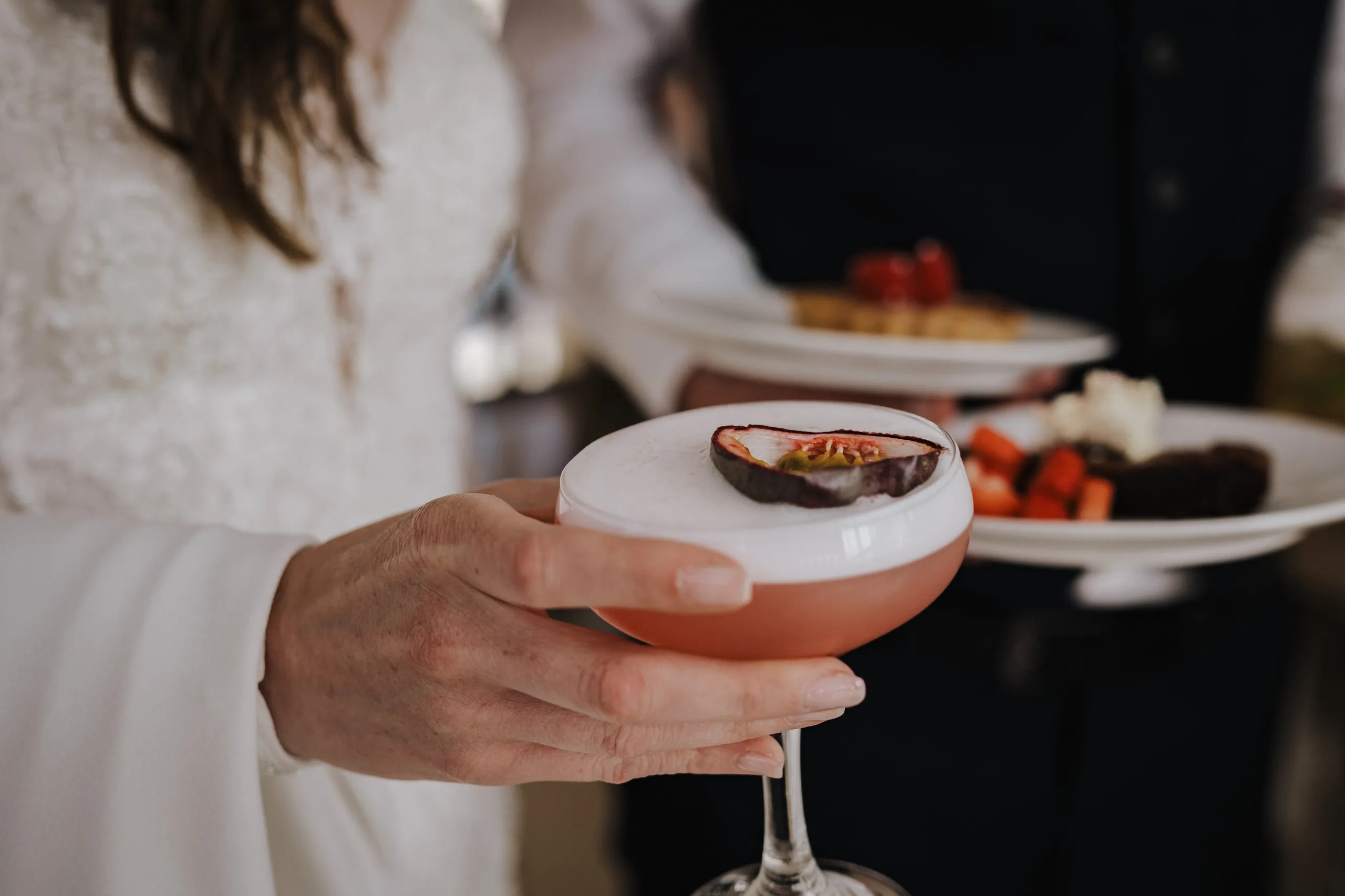 Person holding a pink cocktail with white foam and a passion fruit garnish, with plates of food in the background