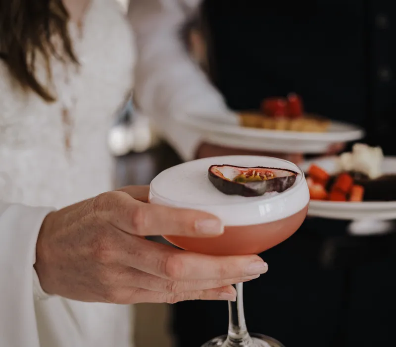 Person holding a pink cocktail with white foam and a passion fruit garnish, with plates of food in the background