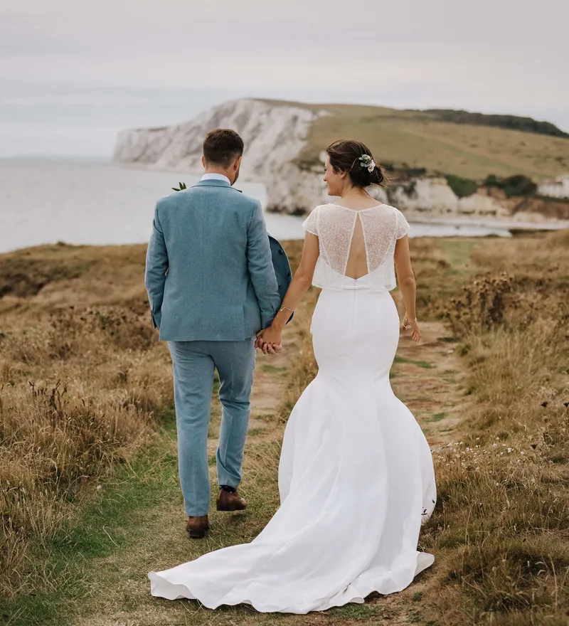 Bride and groom holding hands while walking along a coastal path with white cliffs in the distance