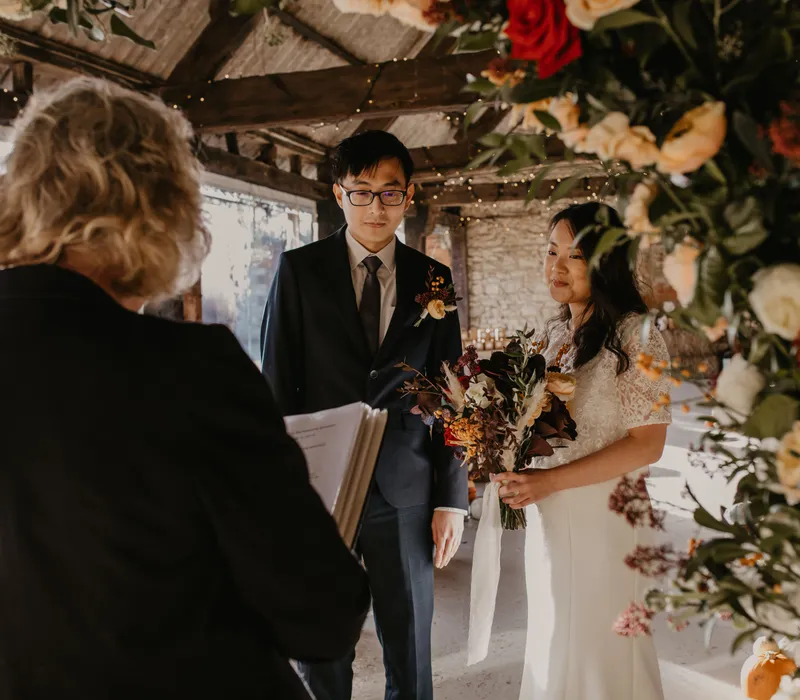 Bride and groom standing in a rustic barn wedding ceremony with floral arch and fairy lights