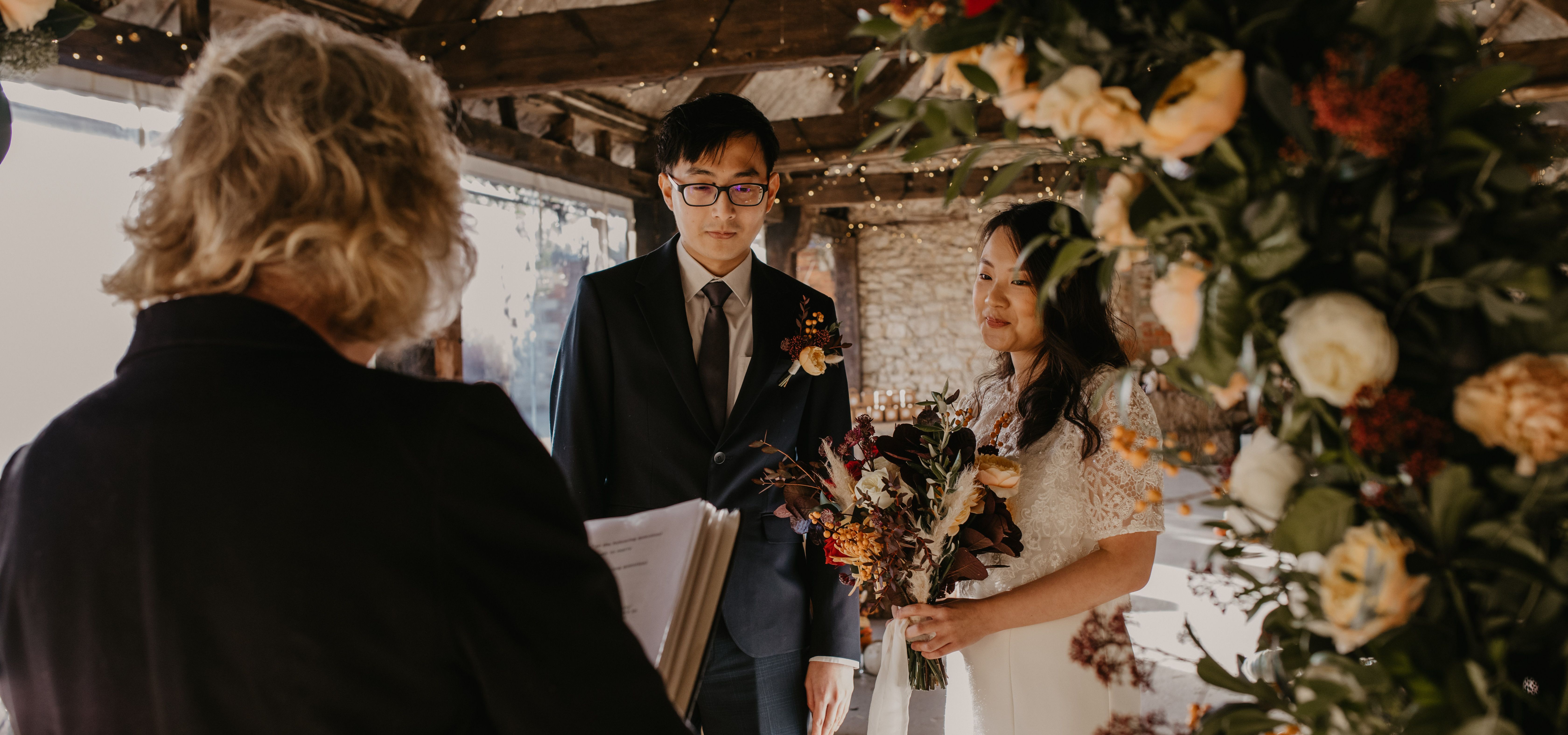 Bride and groom standing in a rustic barn wedding ceremony with floral arch and fairy lights